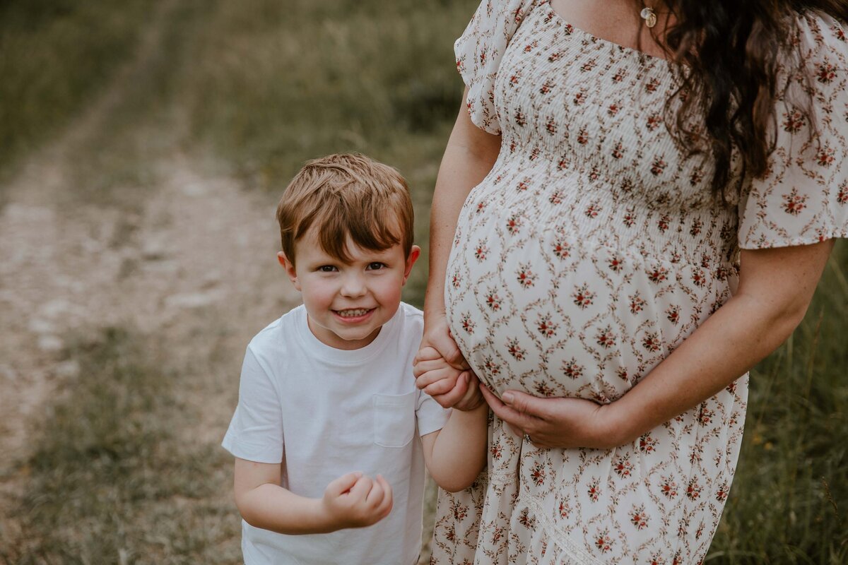Outdoor maternity photo shoot in Exeter, Ontario. Image is a close up of mom's baby bump, with her left hand touching her belly. He is holding her toddler's hand with her left hand and he is smiling at the camera.
