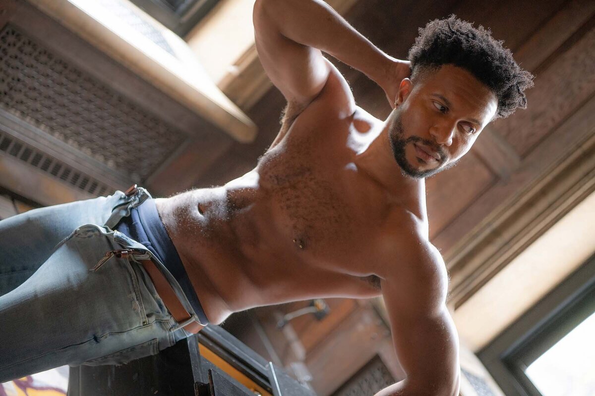 A muscular man with a contemplative expression stands shirtless, wearing jeans, in a sunlit room. He holds his neck, creating a relaxed, introspective tone.