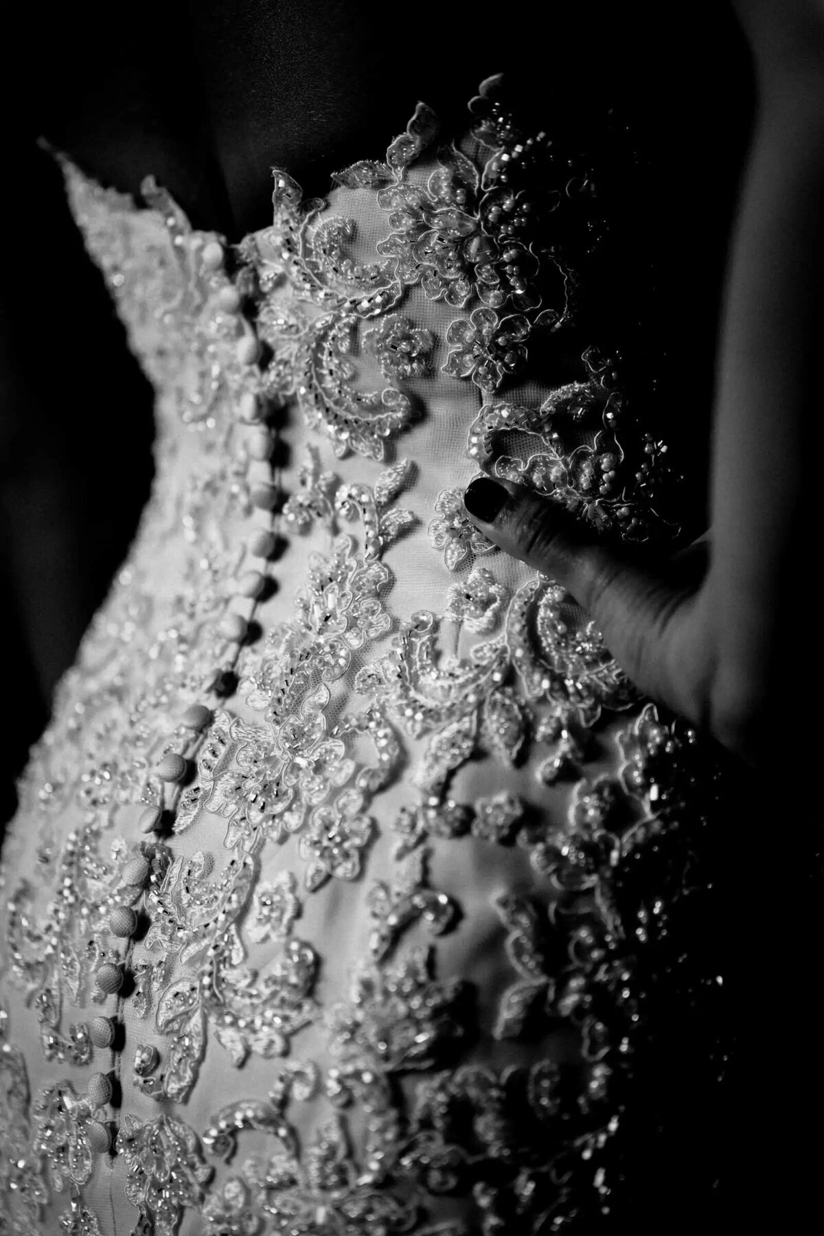 An intimate detail shot of a bride's wedding dress, focusing on the intricate lace and button details, set against a soft-focus black and white background.