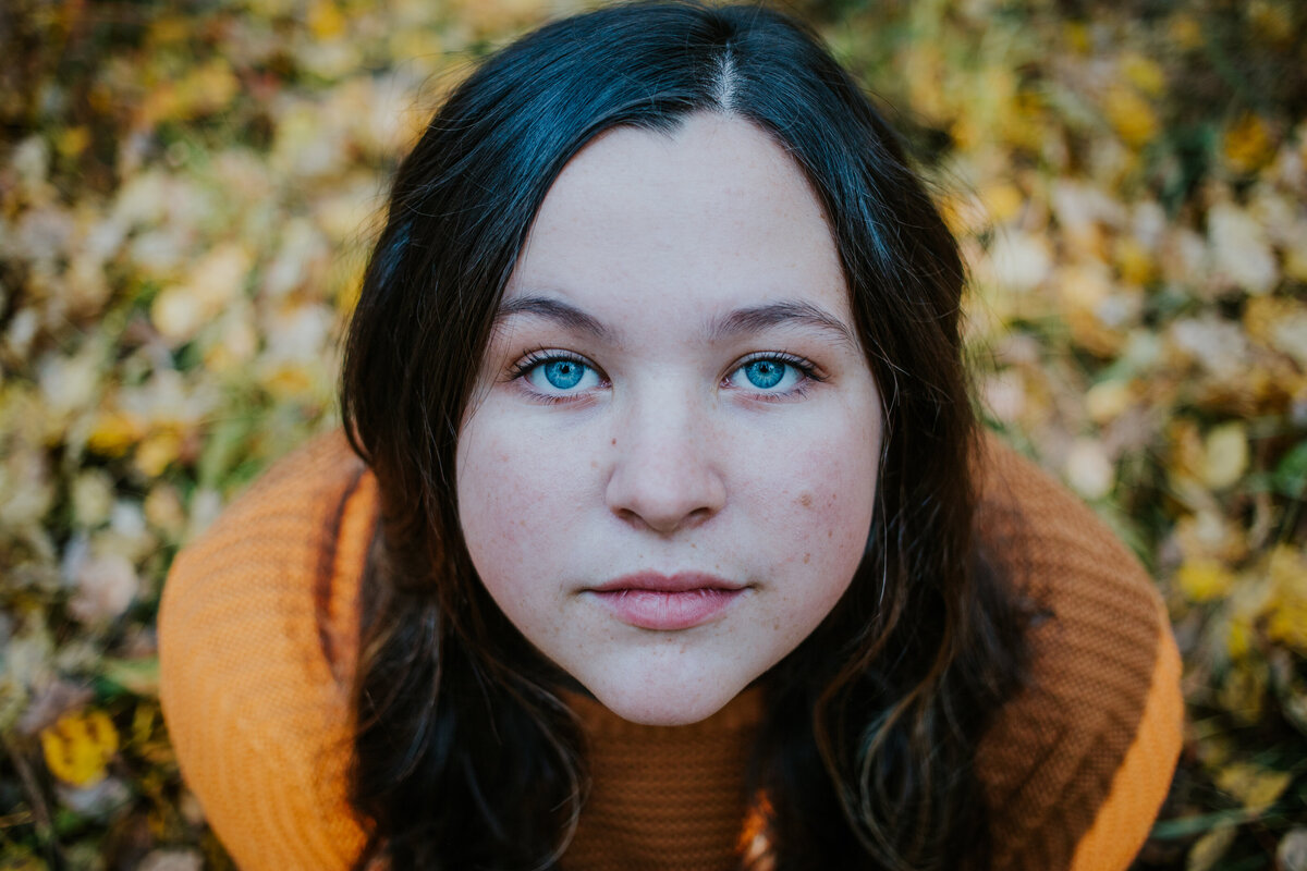 Young woman with bright blue eyes looks into camera while golden leaves surround her.
