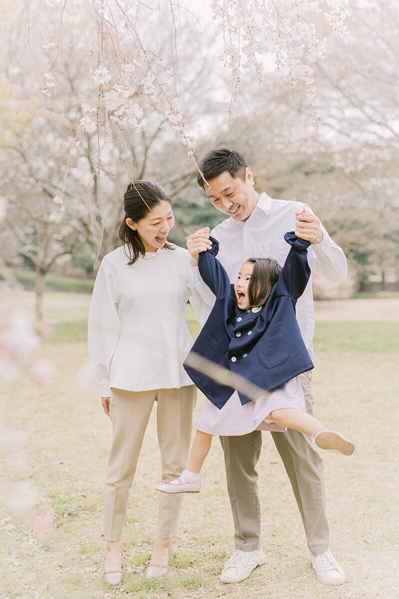 3歳女の子の入園記念の撮影で、桜の木の横でママとパパの3人で撮影。女の子はジャンプをしてパパママと目を合わせて嬉しそうにしている、とても可愛らしくオシャレな家族写真。相模原市でYukika Ishikawa Photographyが撮影。