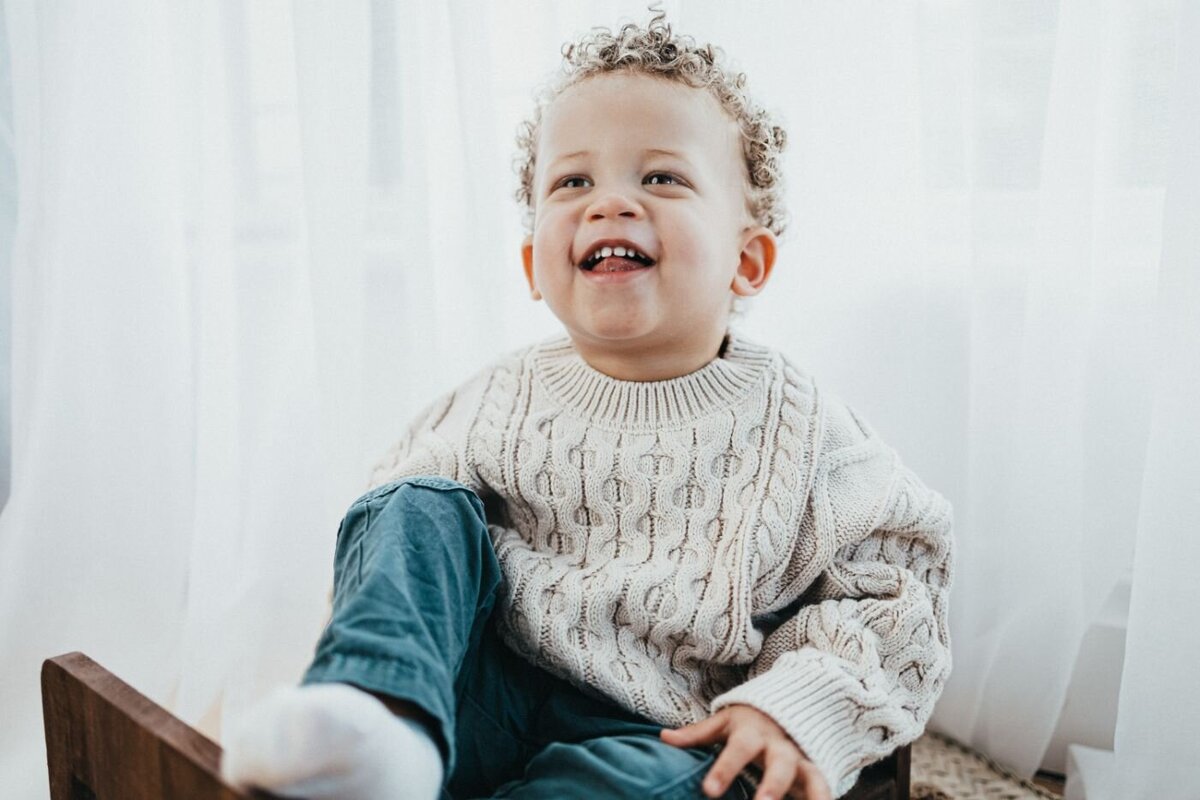 A joyful toddler with curly blonde hair and a wide smile sits in a wooden chair near a bright window with sheer curtain
