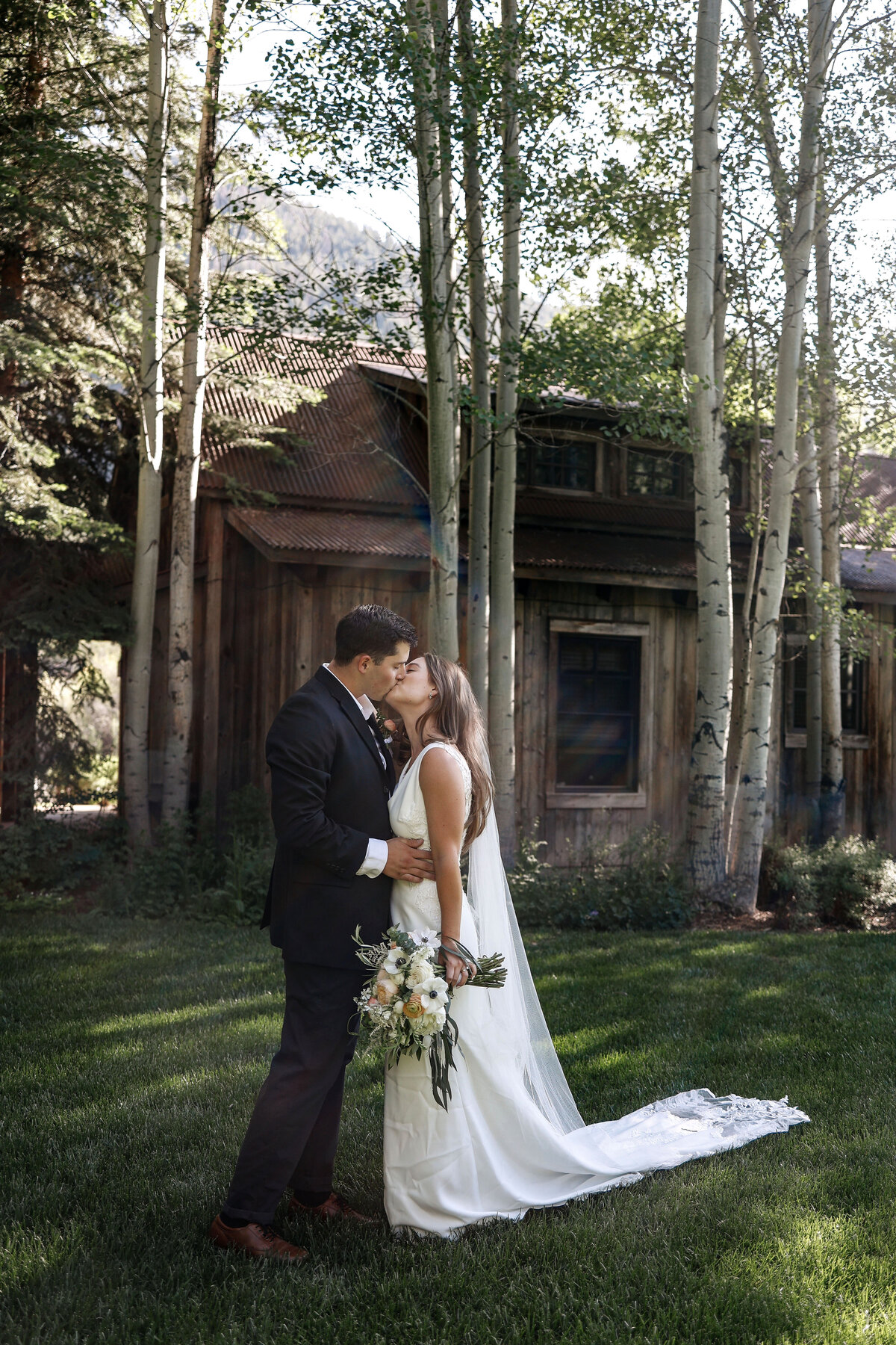 Young bride and groom kiss in front of a rustic barn in Aspen, Colorado,