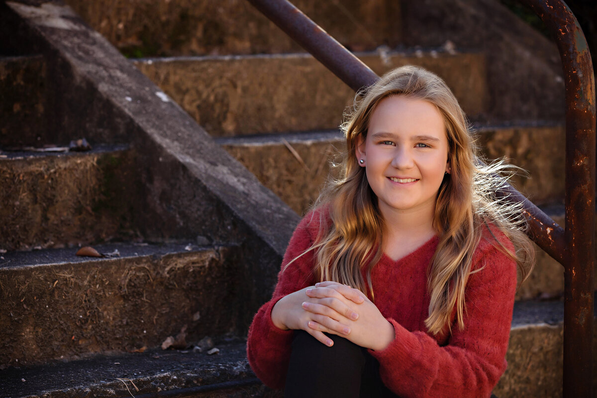 Sweet middle school girl in brick red sweater and black pants sitting on concrete staircase with rust‑colored railings at Camp North End Charlotte NC