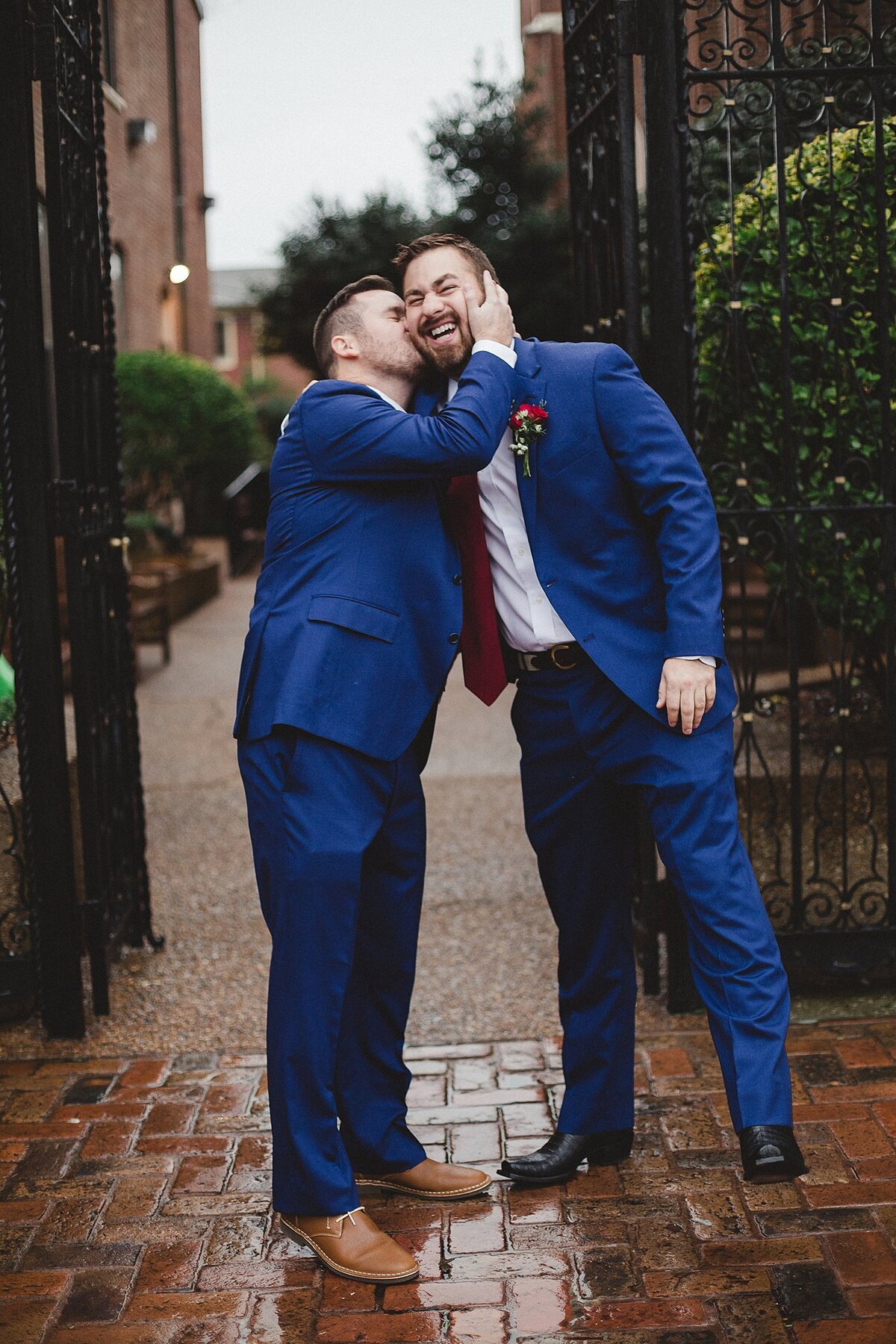 Groom and best man both wearing blue suits with red ties get silly as the best man kisses the groom on the cheek in front of a Nashville church.
