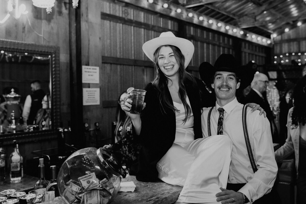 bride and groom wearing cowboy hats sitting on the bar of their west michigan barn wedding reception