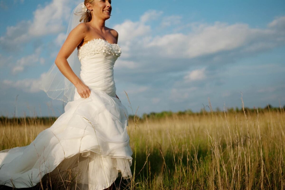 a bride holding up her wedding dress runs through a field of tall grass