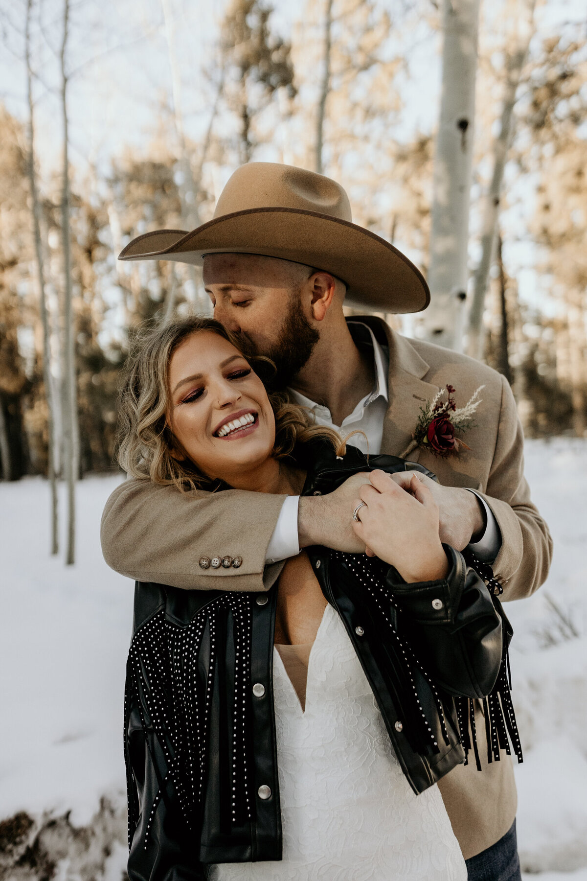 Groom in a cowboy hat hugging bride in the snow