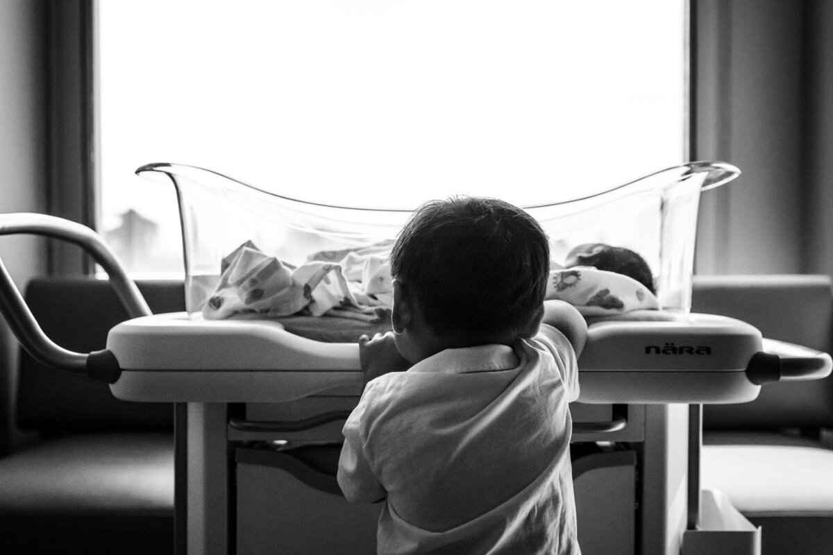 back of toddler brother's head who is looking into hospital bassinet at newborn sister