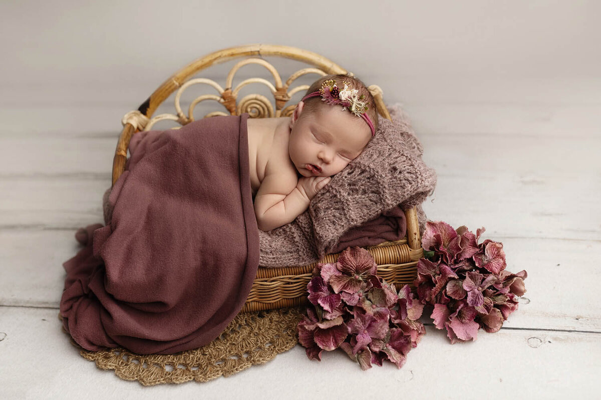 A newborn baby sleeps on a wicker bench under a purple blanket with matching flowers