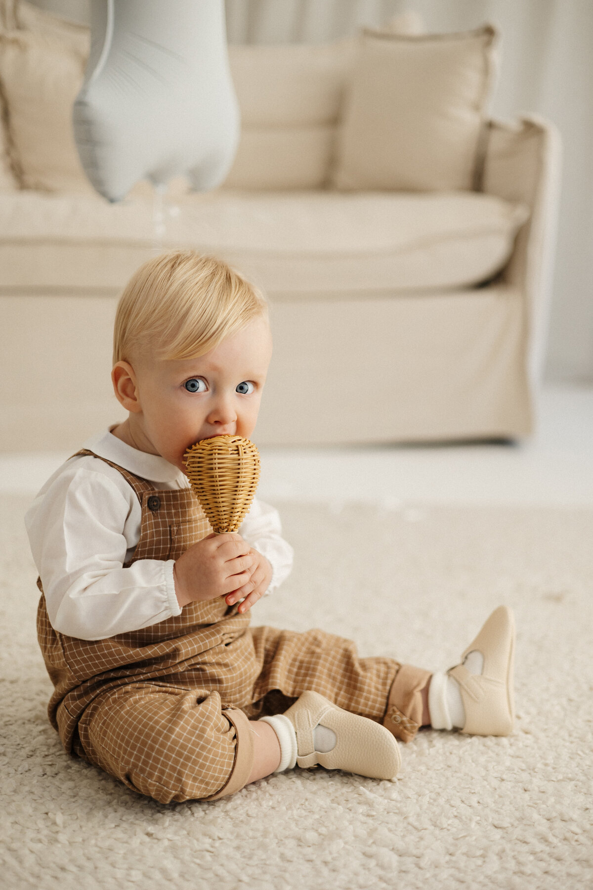 a baby holding a cane rattle and looking at the camera with his big eyes and sitting on the mat in lucy janes photography studio