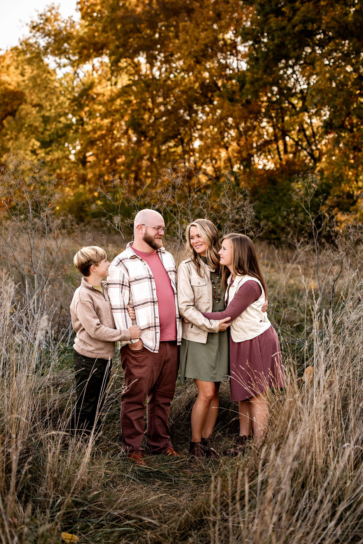 A family of four stands in a field surrounded by tall grass and autumn trees, captured elegantly by a Bloomington family photographer. They are smiling and dressed in casual fall attire. The children are beside the adults, who are embracing each other warmly under the setting sun's glow.