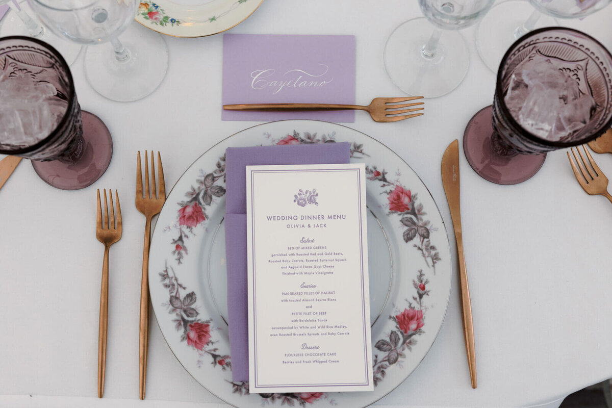 A purple wedding menu card on top of an elegant plate with wine glasses and bronze cutleries around it, at The Ausable Club, NY.