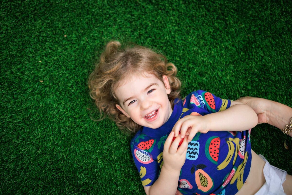 Overhead photo of a toddler boy lying on grass looking up and smiling.