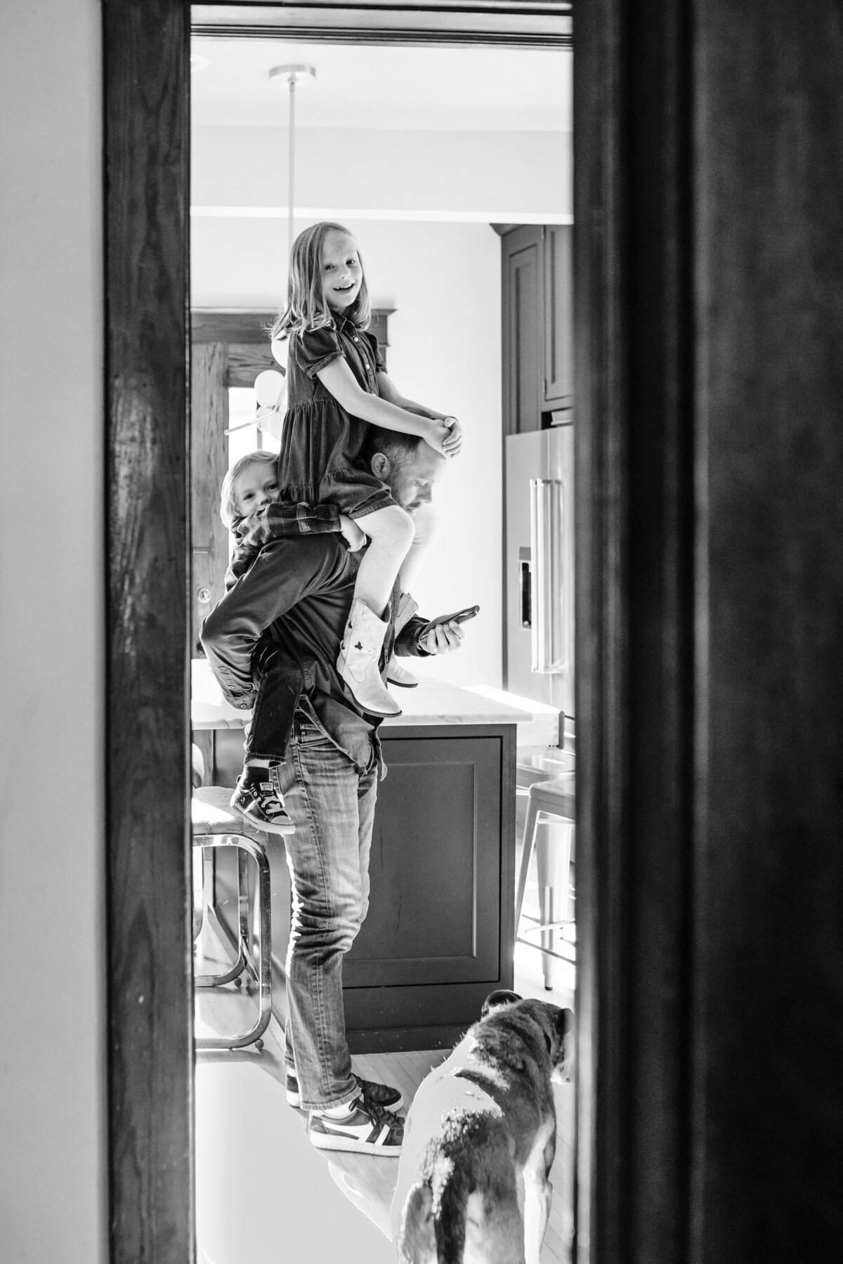 A black and white photo showing a man holding two young children, one on his shoulders and the other on his back, in a warm kitchen setting. A dog is also visible at the bottom, near the man's legs.