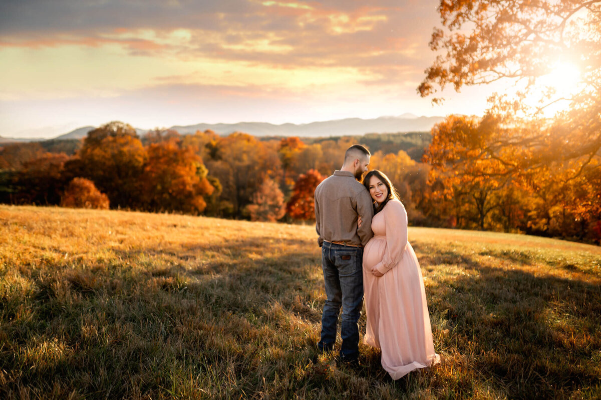 An adorable expecting mama in a long pink dress leans on her husband while standing in a field at sunset