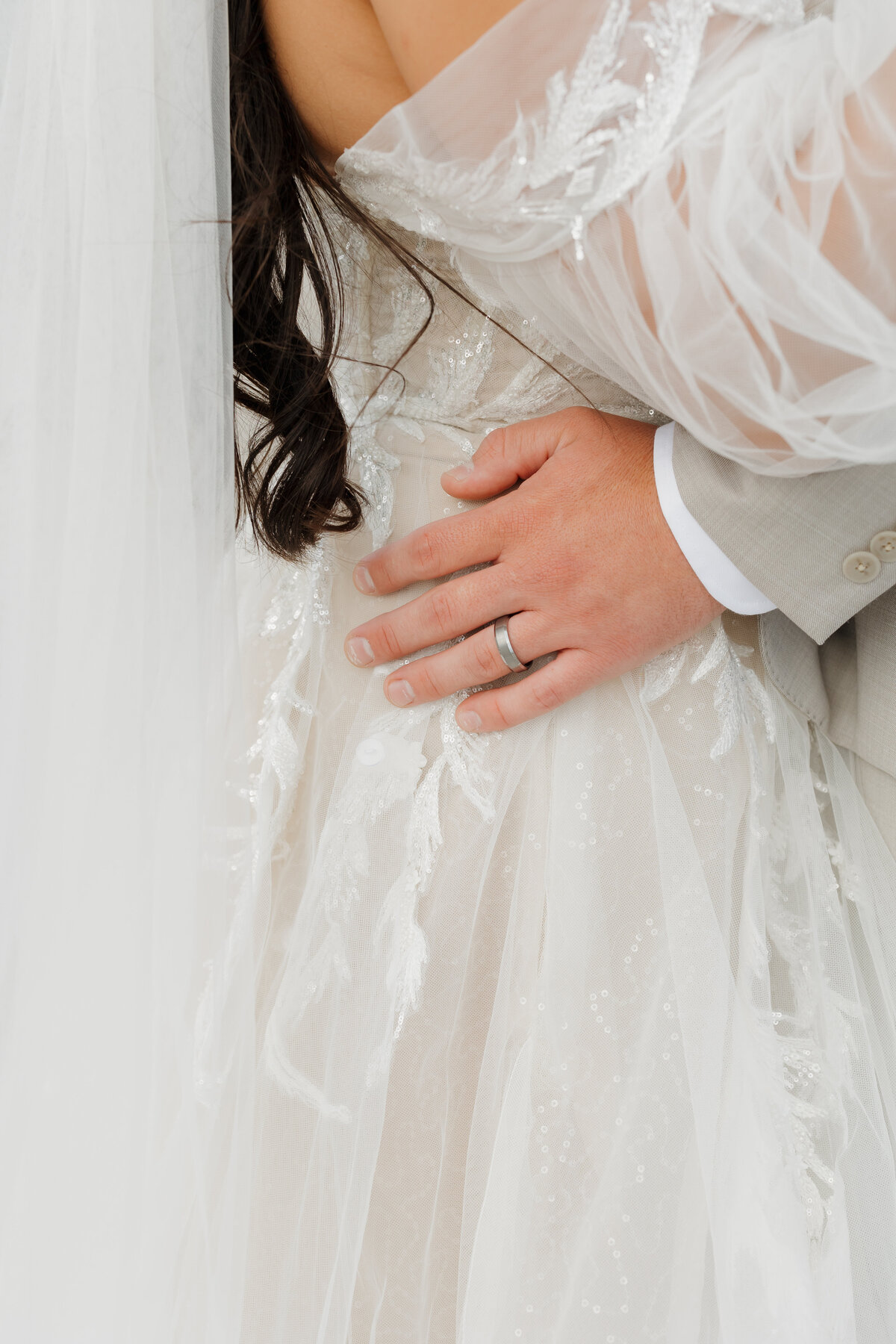 Close-up of a couple embracing on their wedding day, captured by a New England elopement photographer. The bride wears a sheer, embellished gown with a veil. The groom, in a light suit, has his hand on her waist, wearing a dark wedding band. The scene exudes elegance and intimacy.