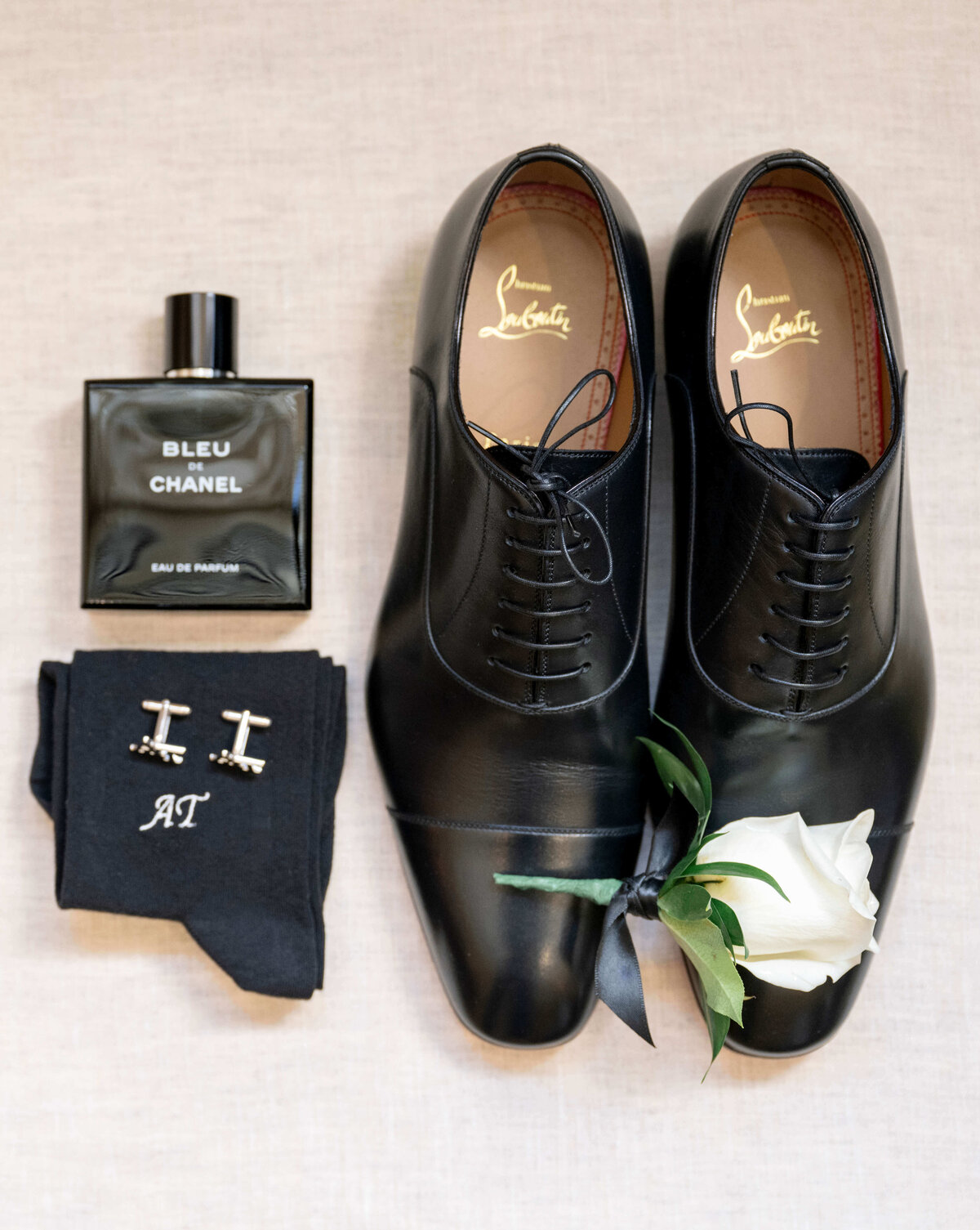 Flat lay of black leather dress shoes, a bottle of Bleu de Chanel cologne, black socks with the initials AT, silver cufflinks, and a white rose boutonniere on a light background.