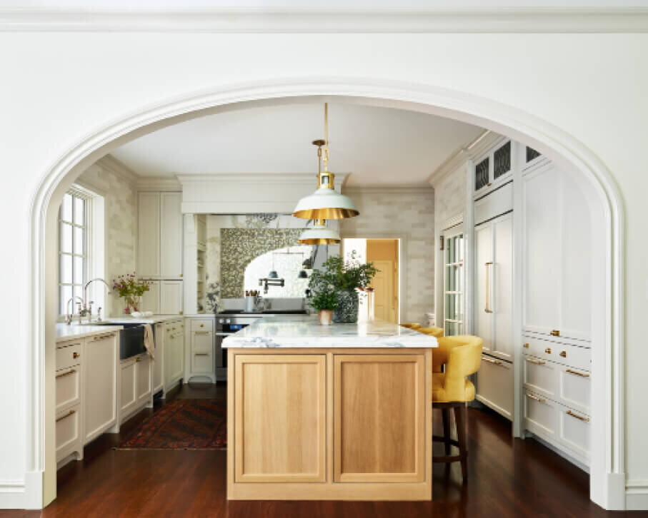 Interior decorators in Boston designed a classic kitchen with a natural oak island, custom yellow stools, white cabinets, brass details, and counter-to-ceiling tile.