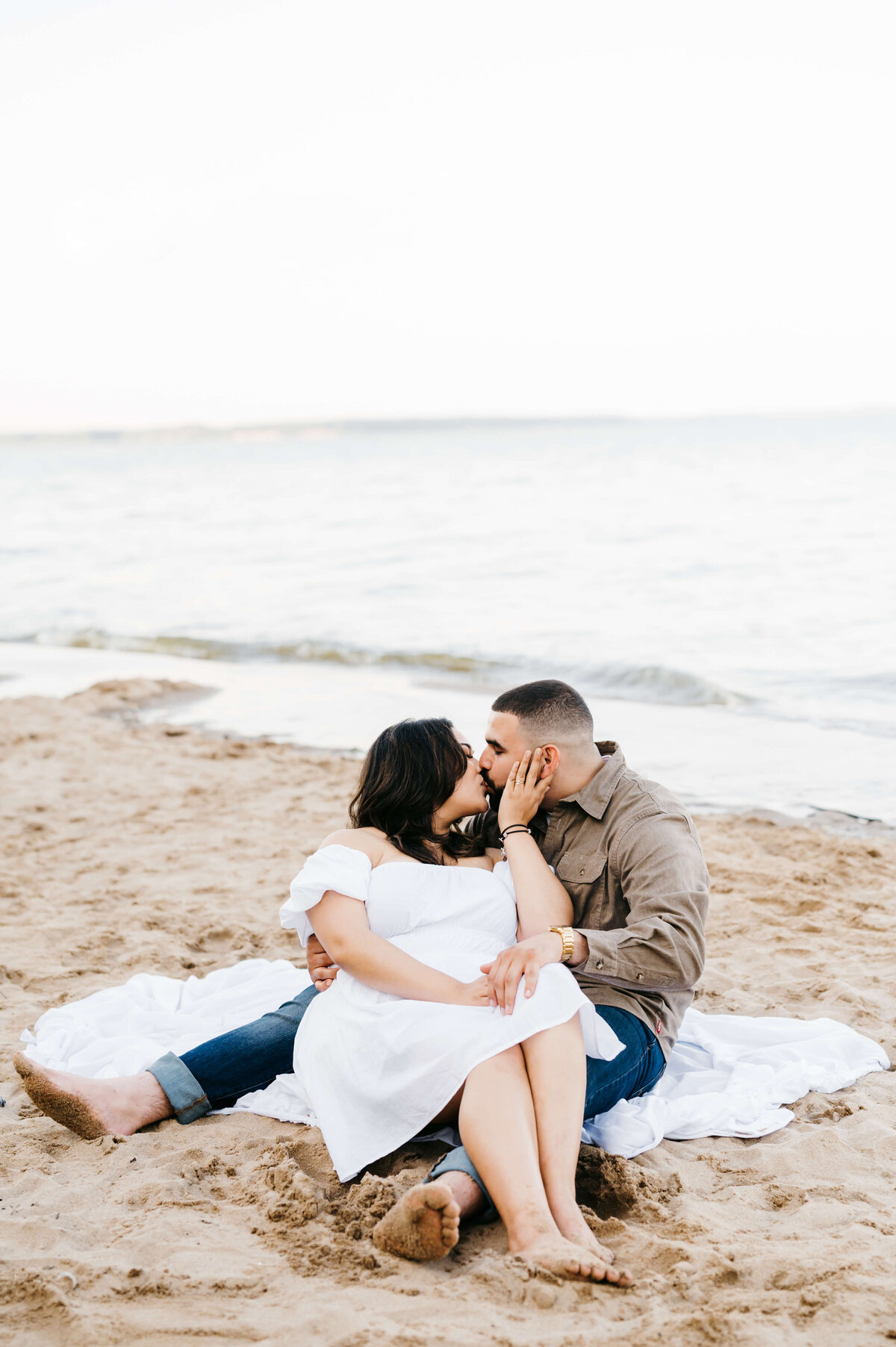 woman wearing a white dress for engagement photo while she sits between a mans legs as he is on a blanket on the beach at woodbridge lake for beach engagement photos