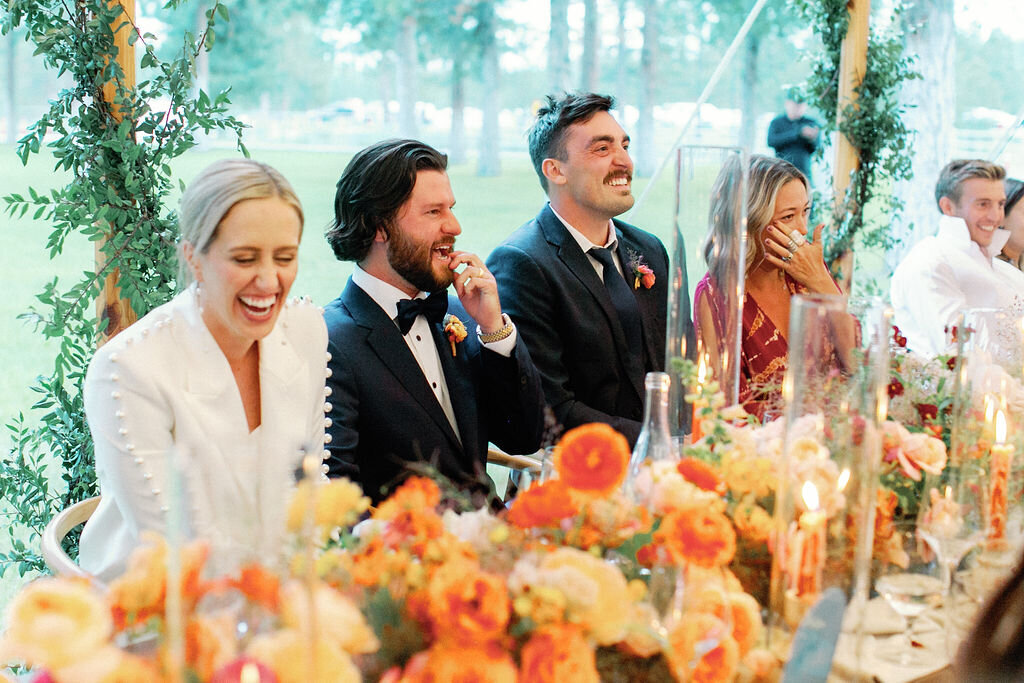 Head table with ombré floral runner in blush, coral, and tangerine tones, with Kelly, Joe, and guests seated during their wedding reception under a sailcloth tent, designed by Summer Robbins Flowers in Bend, Oregon