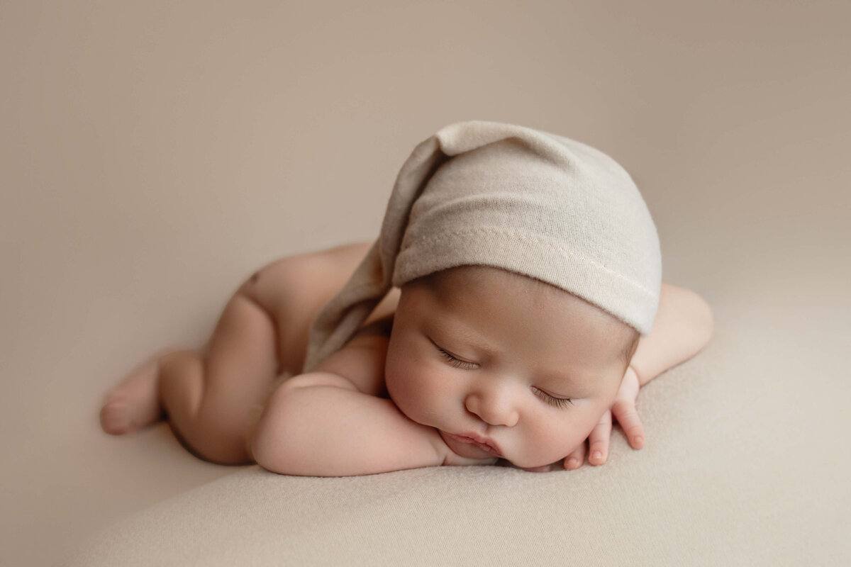 A newborn baby sleeps in froggy pose in a beige sleep cap in a studio