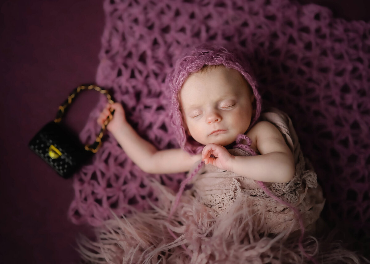 An image from a Pella Newborn Photography session of a baby sleeping in a knit bonnet while holding a tiny black purse