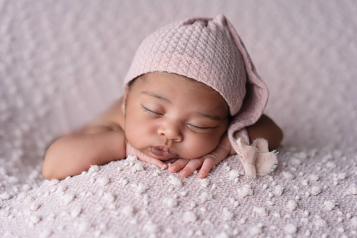 Newborn baby girl sleeping for newborn photoshoot with her hands folded under her chin. She is wearing a long pink knit hat. Captured by Bryn Mwar Philadelphia and Main Line photographer Liza Nicole.