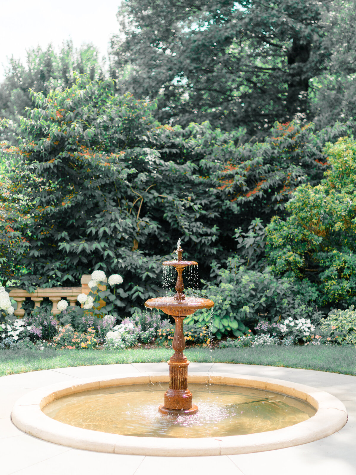 A romantic tiered fountain flows gently in the formal garden of Cairnwood Estate, surrounded by lush greenery and blooming florals. This serene scene captures the estate’s timeless elegance, photographed by Elizabeth Kane Photography, a Luxury Philadelphia Wedding Photographer.