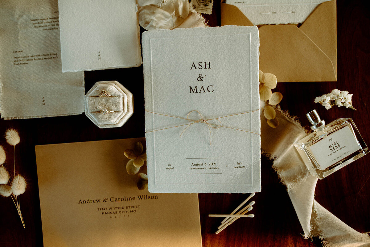 White wedding invitations with black serif text tied with string on a table with tan envelopes and rings