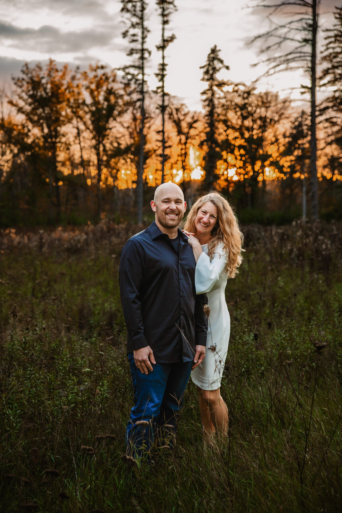 man and woman smile while woman rests her hands on mans shoulders while sun sets through the trees behind them