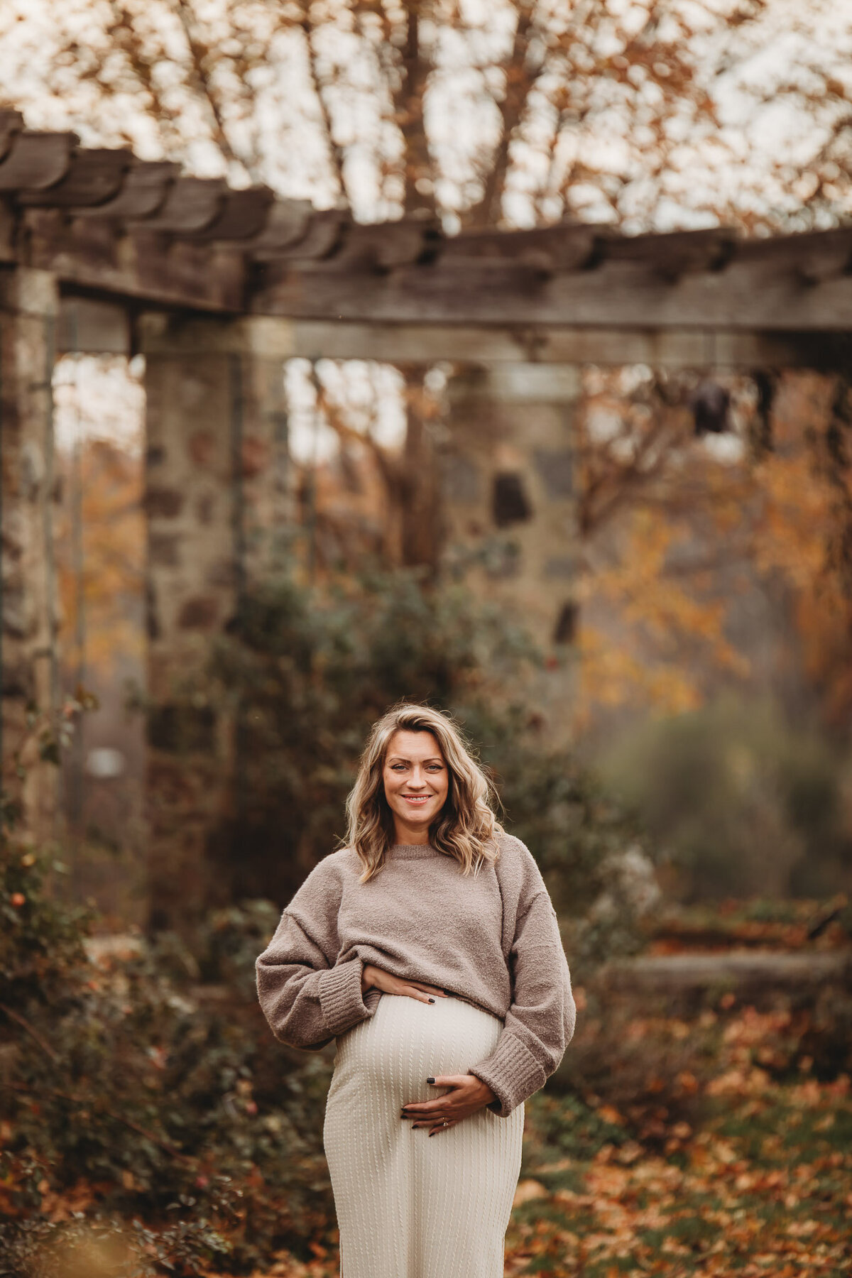 Pregnant mom standing outdoors in the fall in a beige outfit smiling at camera while holding her pregnant belly