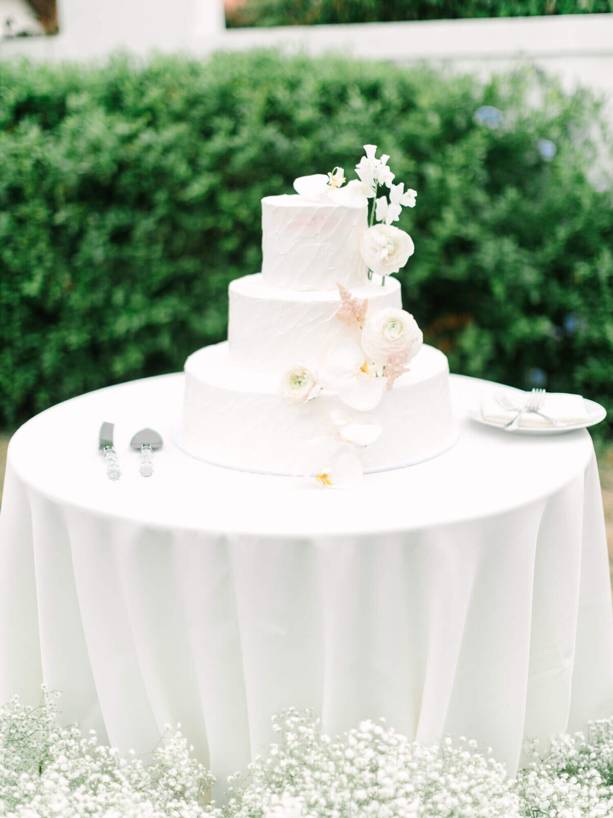 Three-tier white wedding cake adorned with delicate white flowers, on a draped table amidst lush greenery, conveying elegance and celebration.