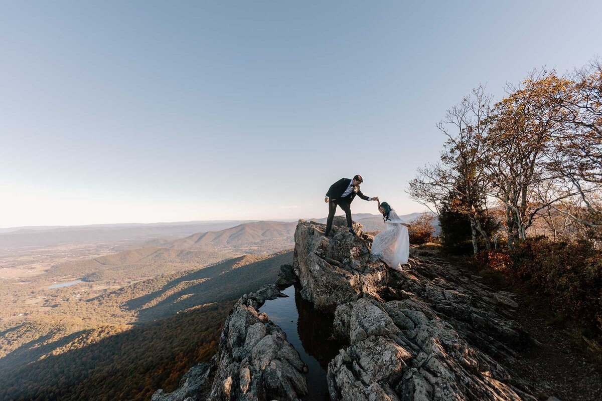 A groom helps his bride to the top of the rock at Little Stonyman in Shenandoah National Park during their elopement