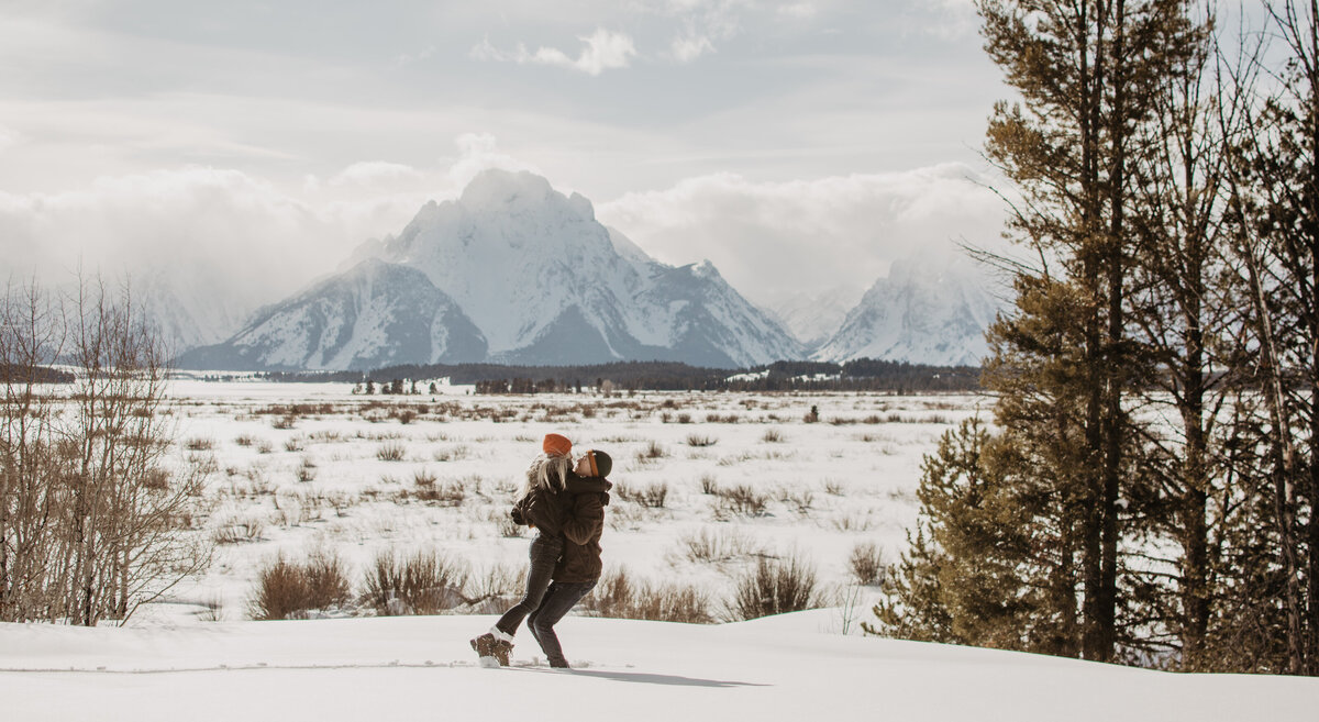 wide angle shot of the Tetons and an engaged couple laughing and holding each other