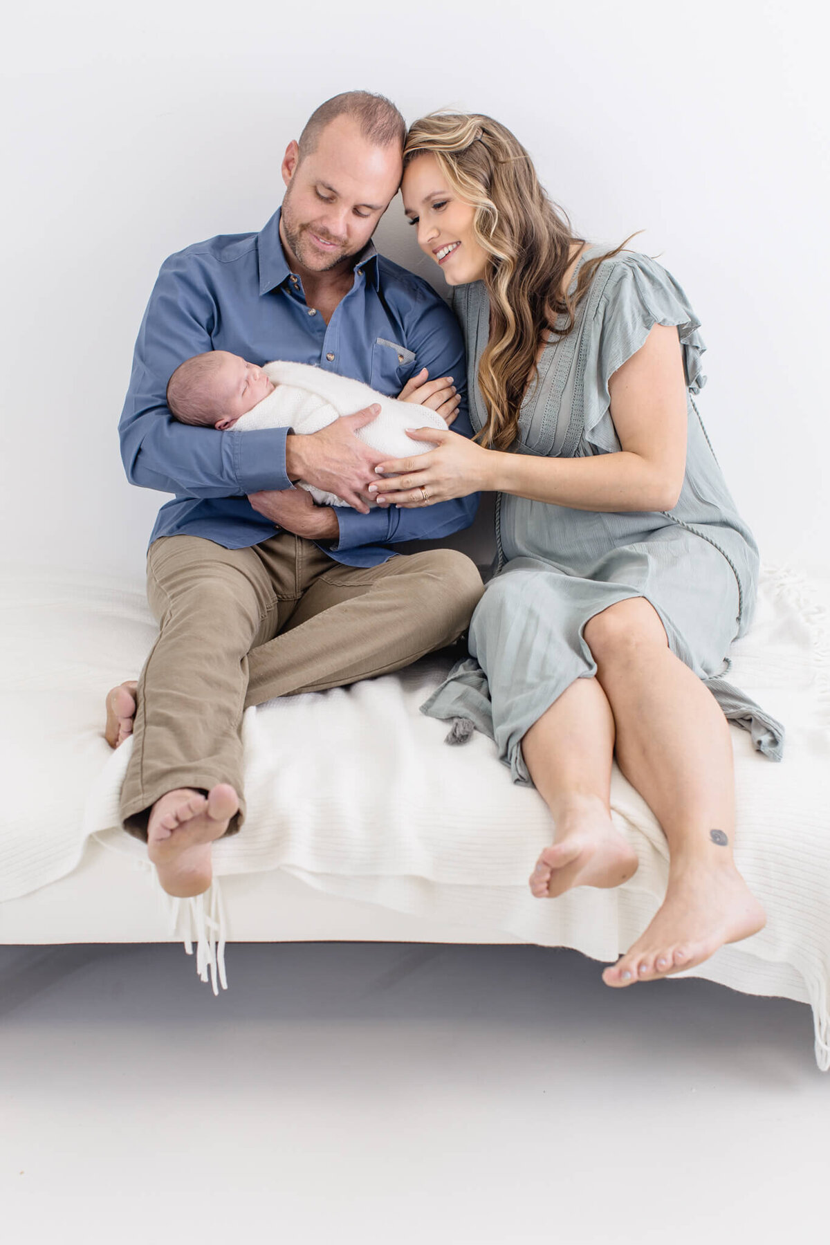 A man and woman sit on a white bed, smiling at a sleeping baby they hold together. The man wears a blue shirt and tan pants, while the woman wears a light blue dress. The setting is bright and airy.