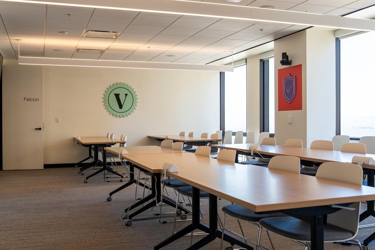 Training or classroom-style workspace with long tables, white chairs, and a mint green V emblem on the wall.