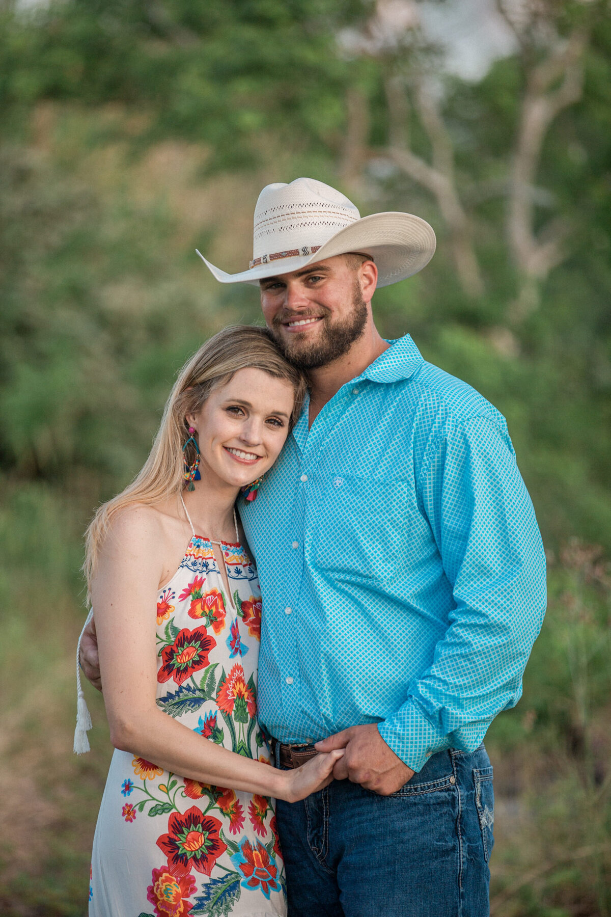 A man dressed as a cowboy his blond fiancee smile for the camera, holding hands