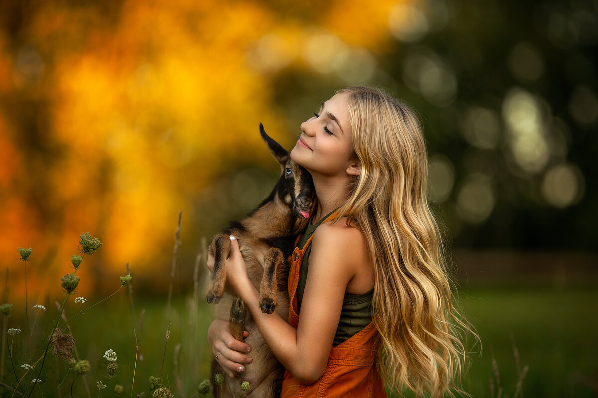 long hair blonde girl holding a goat with a pink tongue in the fall.