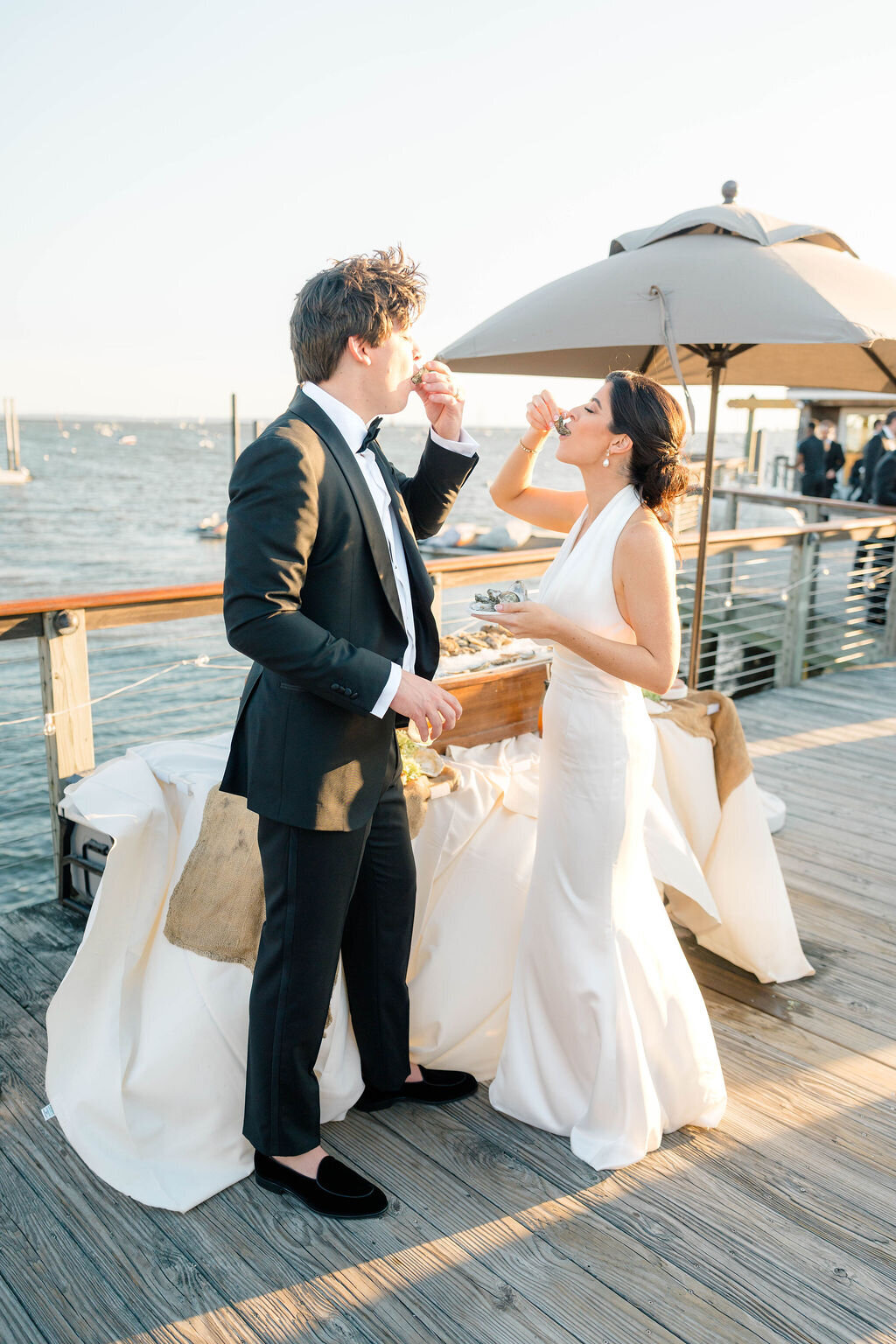 Wedding Photographer RI | A couple in formal attire stands by a waterfront deck, toasting with drinks. The setting sun casts a warm glow.