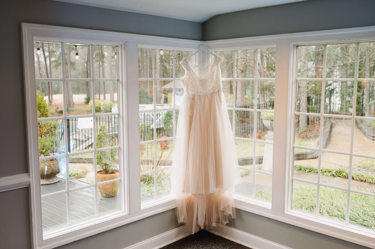 bridal photo of wedding dress hanging in a window in brandermill country club witha lush garden showing through the window