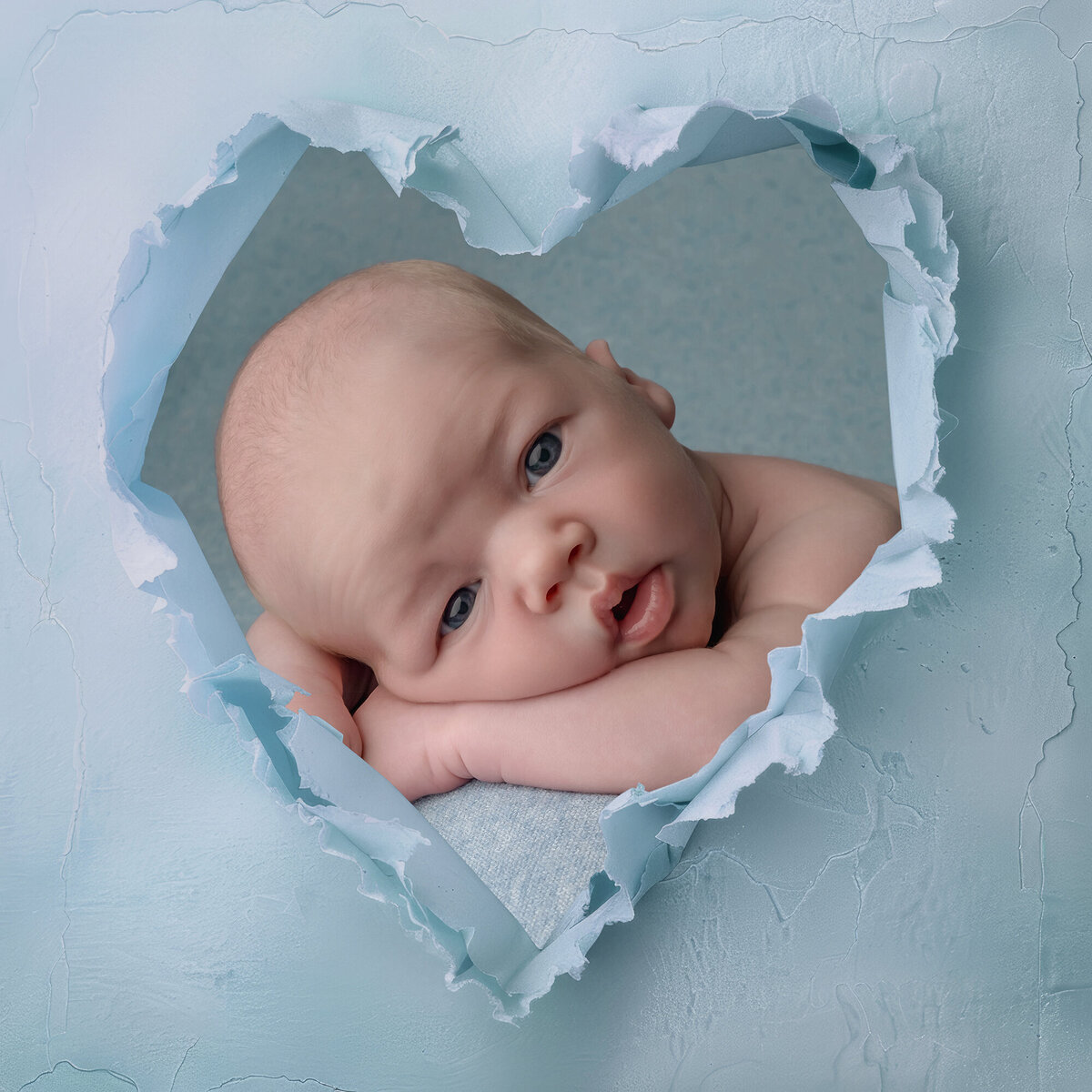 Newborn boy lays behind a sheet of paper with a heart shape ripped in it