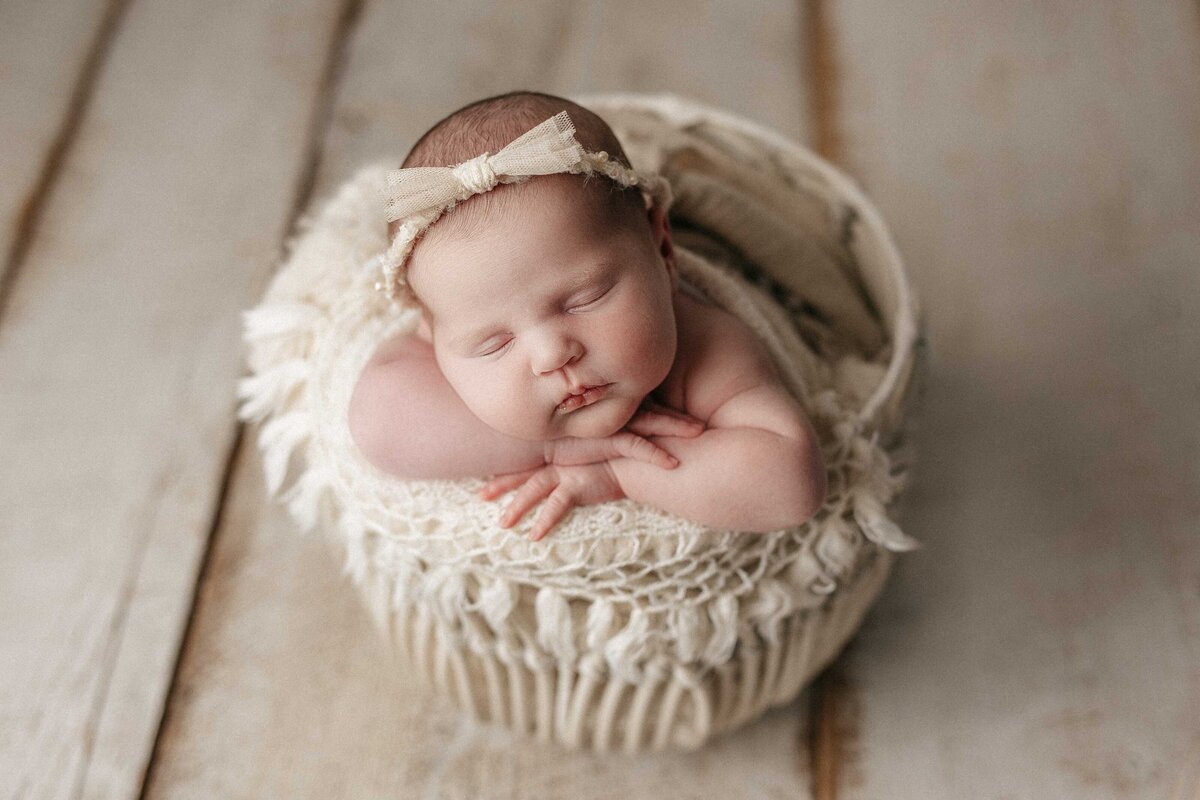 Studio newborn photography - baby sleeping in a basket with a soft blanket that has delicate tassels. Basket is on a wood floor. Baby's hands resting under her chin.