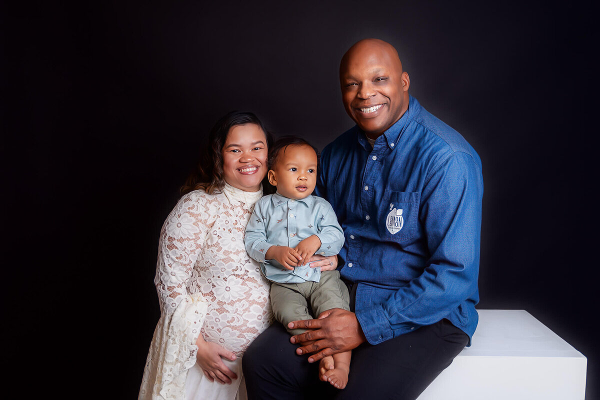 A man and pregnant woman sit on a white box, holding their baby and sharing a joyful moment together.