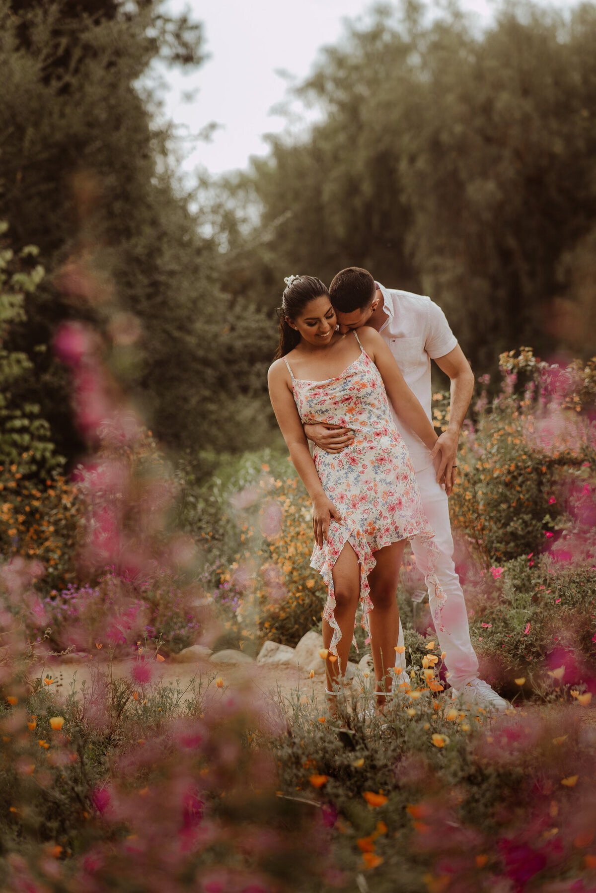 a couple in a flower garden, man hugging her from behind, kissing her shoulder 