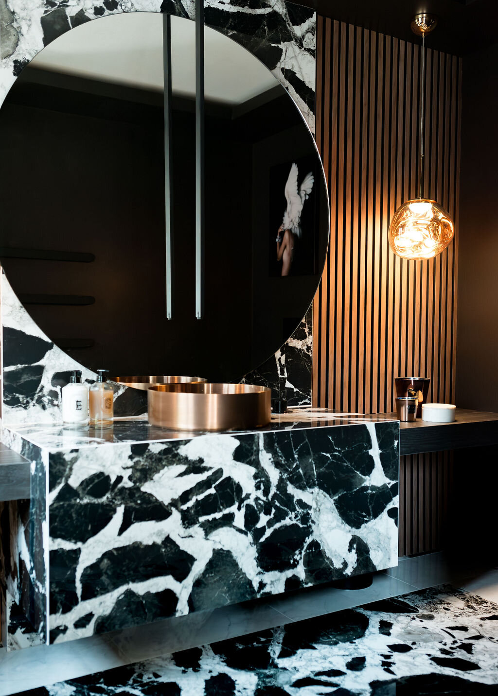 A modern bathroom with a large round mirror, black and white marble sink and countertop, wooden slatted wall, and pendant light fixture.