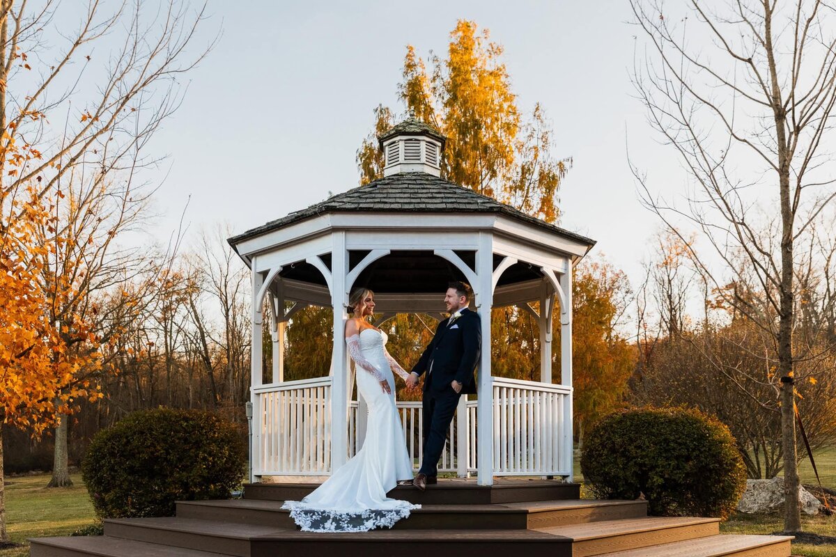A bride and groom stand on the steps of a white gazebo at sunset, holding hands and smiling warmly at each other. The bride’s off-the-shoulder gown features a long lace train that cascades down the stairs, while autumn trees with golden leaves frame the scene in the background.