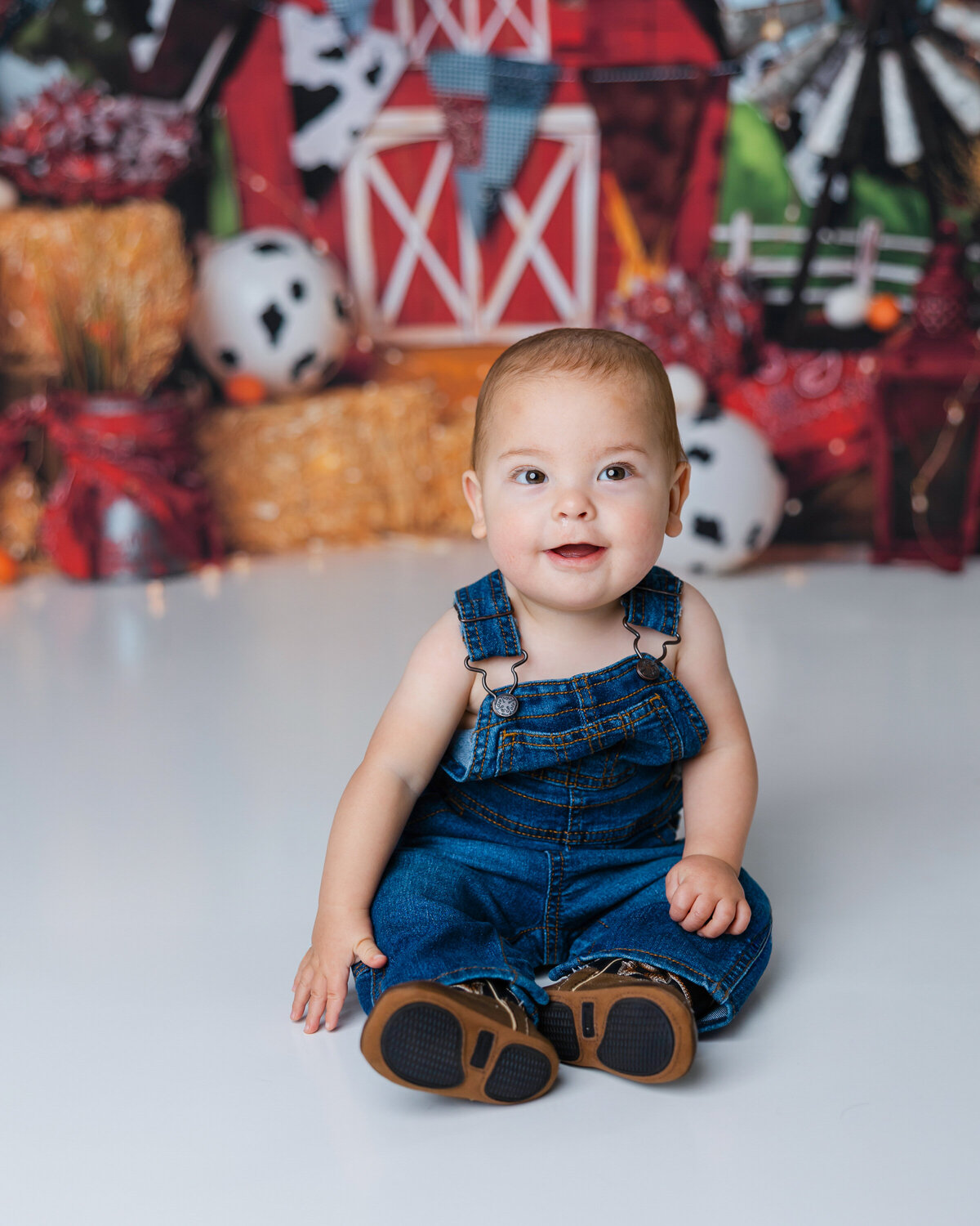 Baby in denim overalls sitting in front of a barn-themed backdrop. Photograph by Yucaipa cake smash photographer Kaitlyn Dawn Photography.