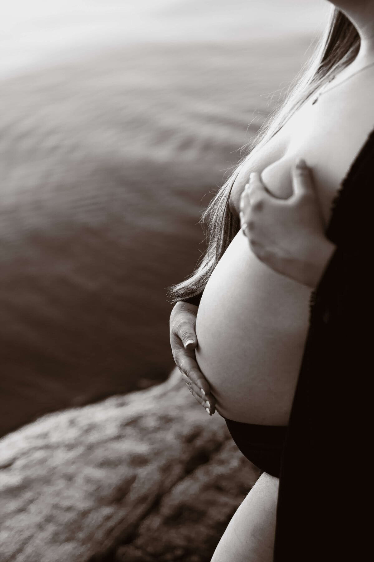 Närbild på gravid mage ståendes på ett berg vid vattnet under en gravidfotografering i Stockholm