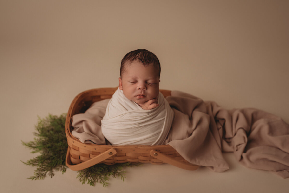 2 week old baby boy at atlanta's best newborn photography studio posed in a cream swaddle wrap sitting in a woven basket with creams and greens